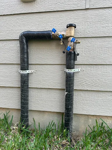 An exposed sprinkler pressure vacuum breaker on insulated pipes against house siding, its blue valve handles turned halfway open